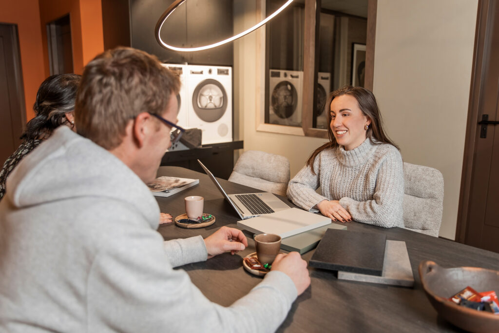 Drie mensen in een moderne vergaderruimte zitten rond een tafel met een laptop, koffiekopjes en materiaalstalen, terwijl een vrouw in een grijze trui glimlachend in gesprek is.