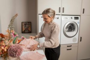 Lachende vrouw in een moderne wasruimte vouwt een zachte, roze handdoek op het aanrecht; op de achtergrond staan ingebouwde wasmachine en droger, terwijl een vaas met bloemen wazig op de voorgrond te zien is.