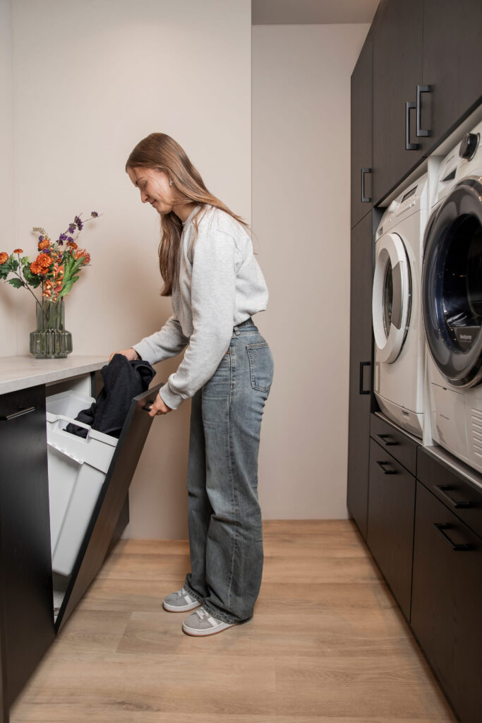 Vrouw legt was in een uittrekbare wasmand in een moderne wasruimte met donkere kasten, houten vloer en wasmachine en droger naast elkaar.